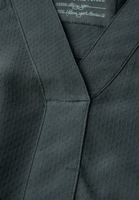 Donkergrijs textiel met een V-hals ontwerp, subtiele perforaties, nette stiksels en een label dat bij de halslijn is geplaatst.