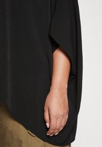 Black top with a textured, lightweight fabric, featuring a loose design and wide sleeves. Hand rests against olive-green trousers.