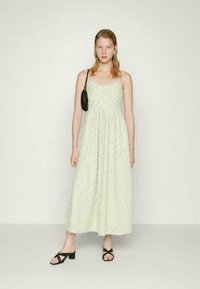 Vestido maxi sem mangas, em verde-claro, com um padrão floral texturizado. Combinado com acessórios pretos e sandálias de salto bloco com a ponta aberta.