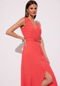 Koronkowa sukienka bez rękawów w kolorze koralowym z dekoltem w kształcie litery V, z paskiem w talii ozdobionym okrągłą metalową klamrą oraz z rozcięciem na udzie po jednej stronie.
