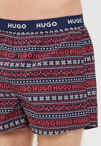 Costum de baie cu un model albastru și roșu, cu un brâu bleumarin care prezintă branding-ul "HUGO". Include desene cu reni și fulgi de zăpadă. Material din bumbac.