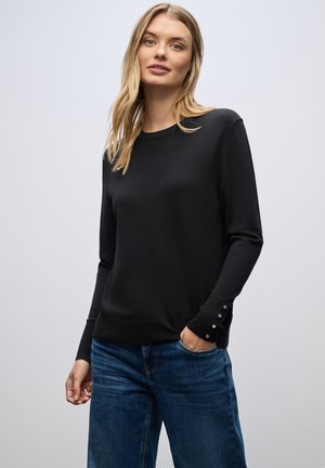 Suéter negro de manga larga en punto suave con cuello redondo y detalles de botones en los puños, combinado con vaqueros de mezclilla azul.