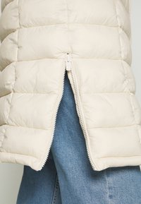 Kremowa kamizelka puchowa z poziomym pikowaniem, z przodem na zamek błyskawiczny i miękką teksturą. Zestawiona z niebieskimi dżinsami, prezentując swobodny styl.