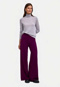 Grauer Rollkragenpullover und weite lila Hosen. Der Pullover ist leicht; die Hosen sind fließend. Das Outfit wird mit schwarzem Schuhwerk kombiniert.