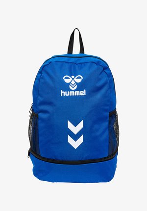 Mochila azul Hummel con logo blanco, dos franjas en forma de cheurón hacia abajo, bolsillos laterales de malla negra y asa superior.