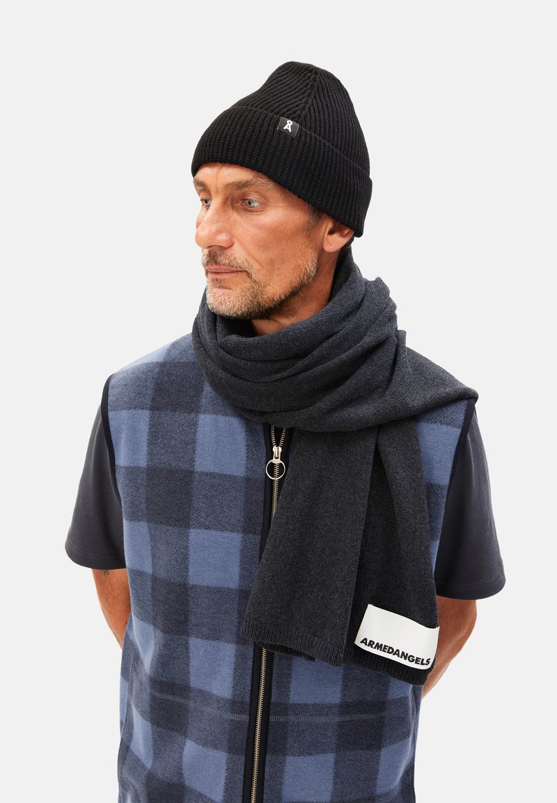 Bonnet en tricots noir, écharpe grise, gilet à carreaux bleus sur une chemise noire. L'écharpe arbore une étiquette blanche avec le logo "ARMEDANGELS".