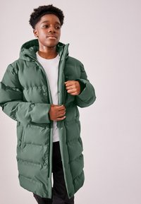 Zeleni puffer kaput s kapuljačom, izrađen od kviltanog, izolacijskog materijala. Ima prednji ZIP zatvarač i kopčanje na dugmad. Model nosi bijelu majicu ispod.