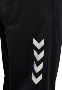 Pantalon de sport noir avec quatre flèches blanches en chevron pointant vers le bas le long de la partie extérieure de la cuisse près de la poche.