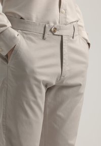 Chinos beiges con cierre de botón, trabillas para cinturón y bolsillos laterales; textura suave y corte ajustado. Color claro y neutro adecuado para varios estilos.