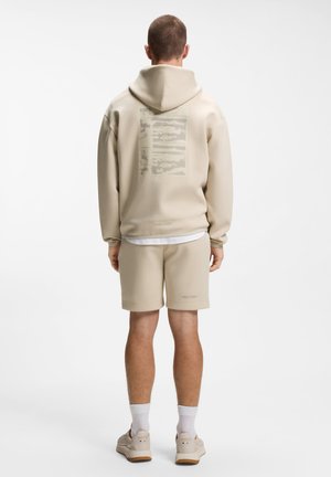 Man standing facing away wearing beige hoodie and matching shorts with white socks and beige sneakers on white background.
