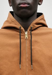 Καφέ ανοιχτό φούτερ με φερμουάρ, κατασκευασμένο από υφασμάτινη υφή, που διαθέτει μαύρες κορδόνες, χρυσό υλικό φερμουάρ και κουκούλα. Ορατή λεπτομερής ραφή.