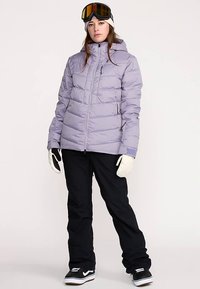 Lichtpaarse gewatteerde jas met ritssluiting, zijzakken en capuchon, gecombineerd met een zwarte broek. Witte handschoenen en skibril maken de outfit compleet.