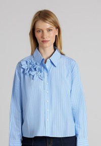 Blusa de rayas azul claro con aplicaciones florales en el hombro izquierdo, que cuenta con un cuello clásico y cierre de botones frontales.