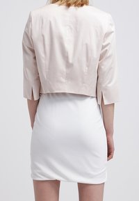 Lichtroze cropped jack met driekwartmouwen, voorzien van zijslitsen, gedragen over een witte aansluitende jurk. Glad, licht glanzend materiaal.