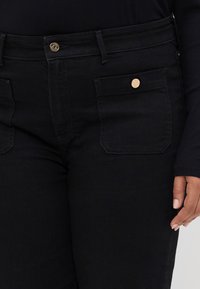 Black denim jeans with a mid-rise waist, two front pockets, and a gold button accent on the waistband. Smooth texture and fitted design.