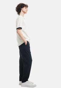 Witte t-shirt met korte mouwen en marineblauwe ribgebreide mouwen, gecombineerd met donkerblauwe broek en witte sneakers, vanuit de zijkant bekeken tegen een effen achtergrond.