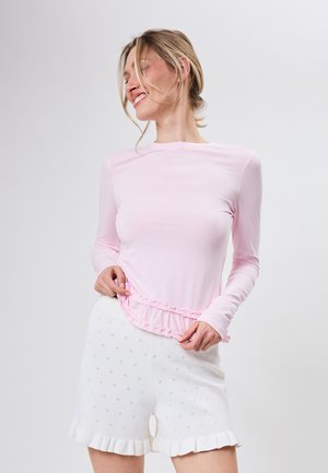 Smilende kvinde i lyserød langærmet top med flæsedetalje og hvide shorts med hulmønster og flæser i kanten, stående mod en ensfarvet baggrund.