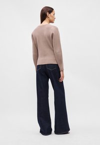 Μπεζ πλεκτή μπλούζα με μακριά μανίκια και στρογγυλή λαιμόκοψη, συνδυασμένη με φαρδιά σκούρα μπλε τζιν παντελόνια, απεικονίζεται από πίσω.