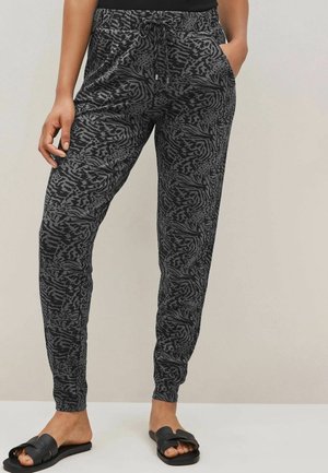 Persoon draagt zwarte en grijze joggingbroek met dierenprint en elastische boorden, zwarte sandalen en een zwart topje, staand tegen een neutrale achtergrond.