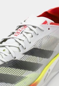 Chaussure de sport blanche avec des accents gris, tige en maille texturée, branding rouge et une semelle jaune-vert vif. Présente des lacets plats et un design épuré.