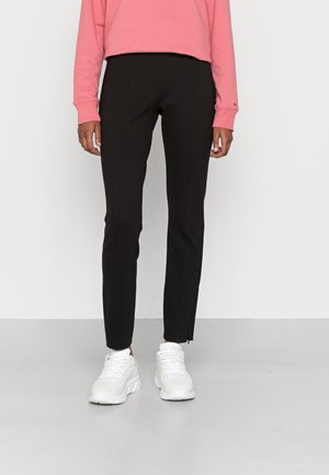 Personne portant un pantalon noir slim, des baskets blanches et un haut rose à manches longues, debout devant un fond clair uni.