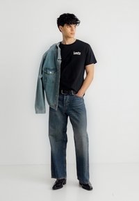 Νέος άνδρας με μαύρο μπλουζάκι Levi's, χαλαρά μπλε τζιν, μαύρα παπούτσια και τζιν μπουφάν ριγμένο στον έναν ώμο, στέκεται δίπλα σε λευκό τοίχο.