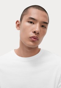 Weißes T-Shirt mit Rundhals, aus weichem Baumwollmaterial, mit entspannter Passform. Das Modell hat kurze Haare und einen neutralen Gesichtsausdruck, vor einem minimalen Hintergrund.