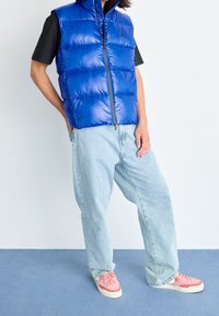 Blå vadderad väst med dragkedja, bärs över en svart t-shirt. Personen har på sig ljusblå jeans och rosa sneakers. Enkel bakgrund.