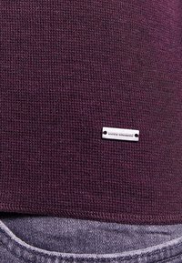 Burgundy texturerad t-shirt med en liten silveretikett som står "BOSS ORANGE" nära fållen, i kombination med ljus denimtyg.