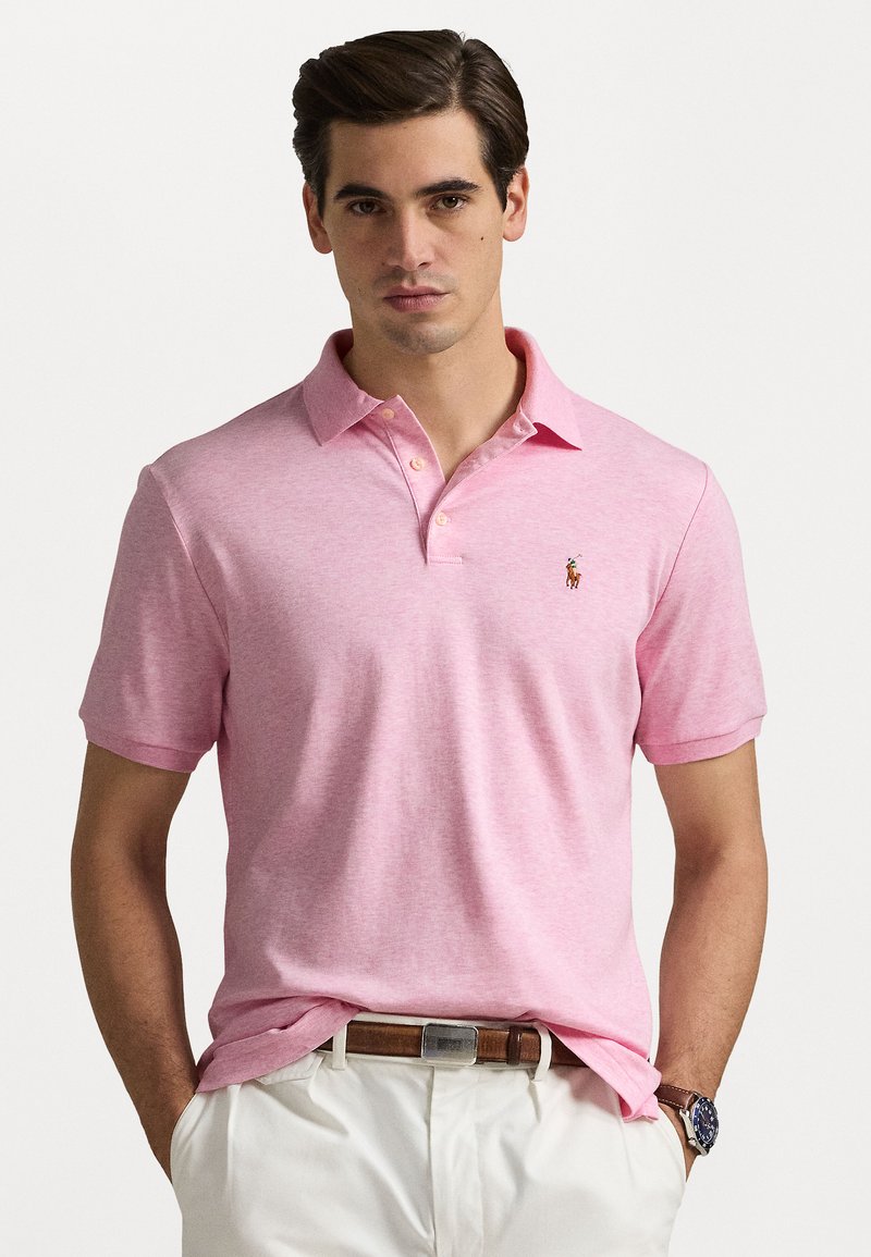 Polo de algodón en rosa claro con cuello clásico, mangas cortas y un pequeño parche de logo en el lado izquierdo del pecho. Combinado con un cinturón marrón.