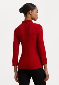 Red, ribbed long-sleeve top with a collar, fitted design, and cuffed sleeves. Smooth texture, hip-length cut, and subtle detailing at the hem.