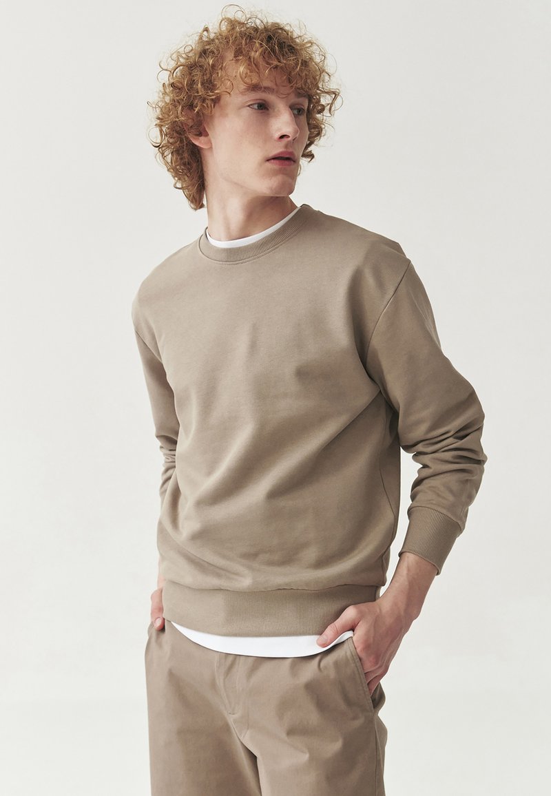 Felpa beige realizzata in morbido tessuto, con collo rotondo, maniche lunghe e polsini a costine. Abbinata a una t-shirt bianca.