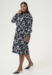 Bloemen jurk in marineblauw met witte bloemen, getailleerde taille, lange mouwen en zijsplitsen. Gecombineerd met zwarte hoge muiltjes.