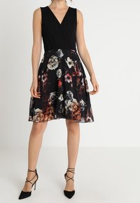 Robe noire sans manches avec un corsage style wrap et un motif floral dans des teintes de rouge, blanc et violet sur une jupe noire. Présente une forme évasée.