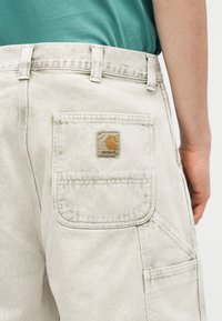 Μπεζ παντελόνια εργασίας Carhartt με τετράγωνη πίσω τσέπη που φέρει το έμβλημα Carhartt, φορεμένα με πράσινο πουκάμισο.