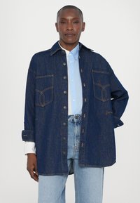 Denim shirt jacket features a dark blue color, two chest pockets with orange stitching, and button closure. Worn over a light blue collared shirt.