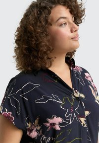 Eine Frau mit lockigem braunem Haar trägt eine dunkelblaue Bluse mit rosa, cremefarbenen und grünen Blumenmustern. Sie schaut leicht nach oben und nach rechts.