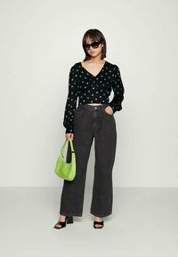 Top preto com bolas brancas e mangas compridas, combinado com jeans escuros de cintura alta. Segura uma pequena carteira verde, usando óculos de sol pretos.