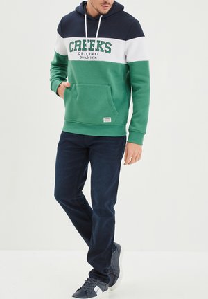 Felpa color-block verde e blu navy con strisce bianche, tasca a marsupio e scritta del logo. Indossata con jeans blu scuro e sneakers grigie.