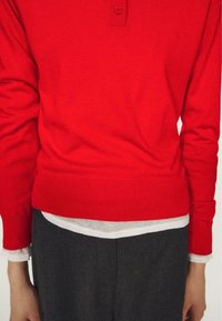 Maglione rosso lavorato a maglia con colletto abbottonato, indossato sopra una camicia bianca. Il maglione ha una fascia aderente in vita e maniche lunghe.