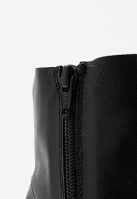 Primer plano de una cremallera de bota de cuero negro con la marca YKK, mostrando cuero texturizado y costuras detalladas sobre un fondo blanco.