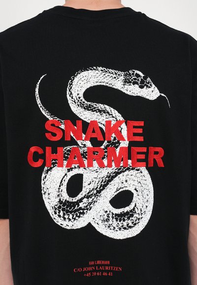 T-shirt noir avec un graphisme de serpent blanc et un texte rouge indiquant "SNAKE CHARMER". Le design a un look audacieux et contrasté.