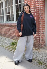 Fekete, oversized kapucnis pulóver kör alakú logóval, világosszürke, bő lábú farmernadrág és fekete papucs. Barna steppelt válltáska. Városi háttér.