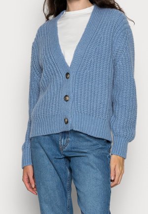 Kobieta w jasnoniebieskim grubym kardiganie na guziki, w białej bluzce i niebieskich dżinsach, stojąca na jednolitym tle.