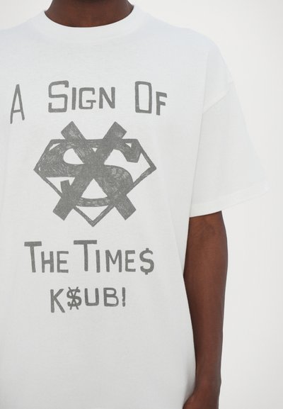 White cotton t-shirt featuring black graphic text: "A Sign Of The Times K$UB!" and a dollar sign inside a diamond shape, with crossed bars.