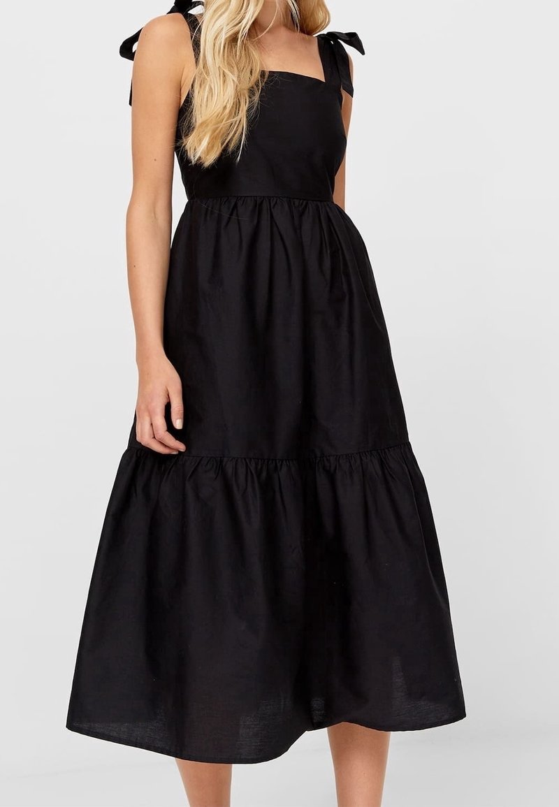 Femme portant une robe midi noire sans manches avec des bretelles nouées en nœud sur les épaules et une jupe à étages.