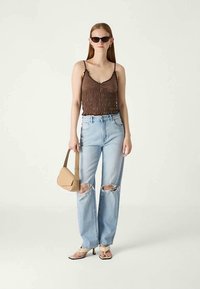 Mulher a usar um top de alças franzido castanho, jeans claros rasgados, mala bege, sandálias de salto e óculos escuros escuros em fundo branco.