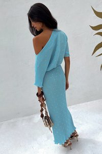 Lichtblauwe off-shoulder jurk met geplooide textuur, versierd met kleine gouden accenten. Hierbij houdt het model een kleine, decoratieve handtas vast.