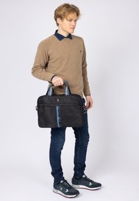Ung mand i beige sweater og jeans, der holder en sort U.S. Polo Assn. mappe med mærkede håndtag mod en hvid baggrund.