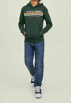 Personne portant un sweat à capuche vert foncé avec le texte "Jack & Jones Athleisure 1990", un jean bleu et des baskets bleu marine et blanches, se tenant avec les mains dans les poches.